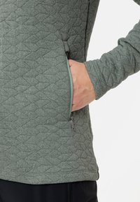 Groene gewatteerde jas met een diagonaal ruitpatroon, een ritszak opzij en lange mouwen met ribboordjes.