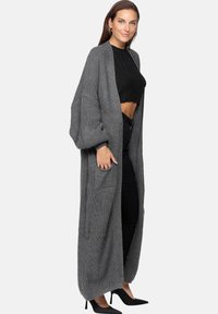 Lungo cardigan grigio a maglia con maniche oversized, apertura frontale e due tasche anteriori, abbinato a un top nero corto e pantaloni neri.