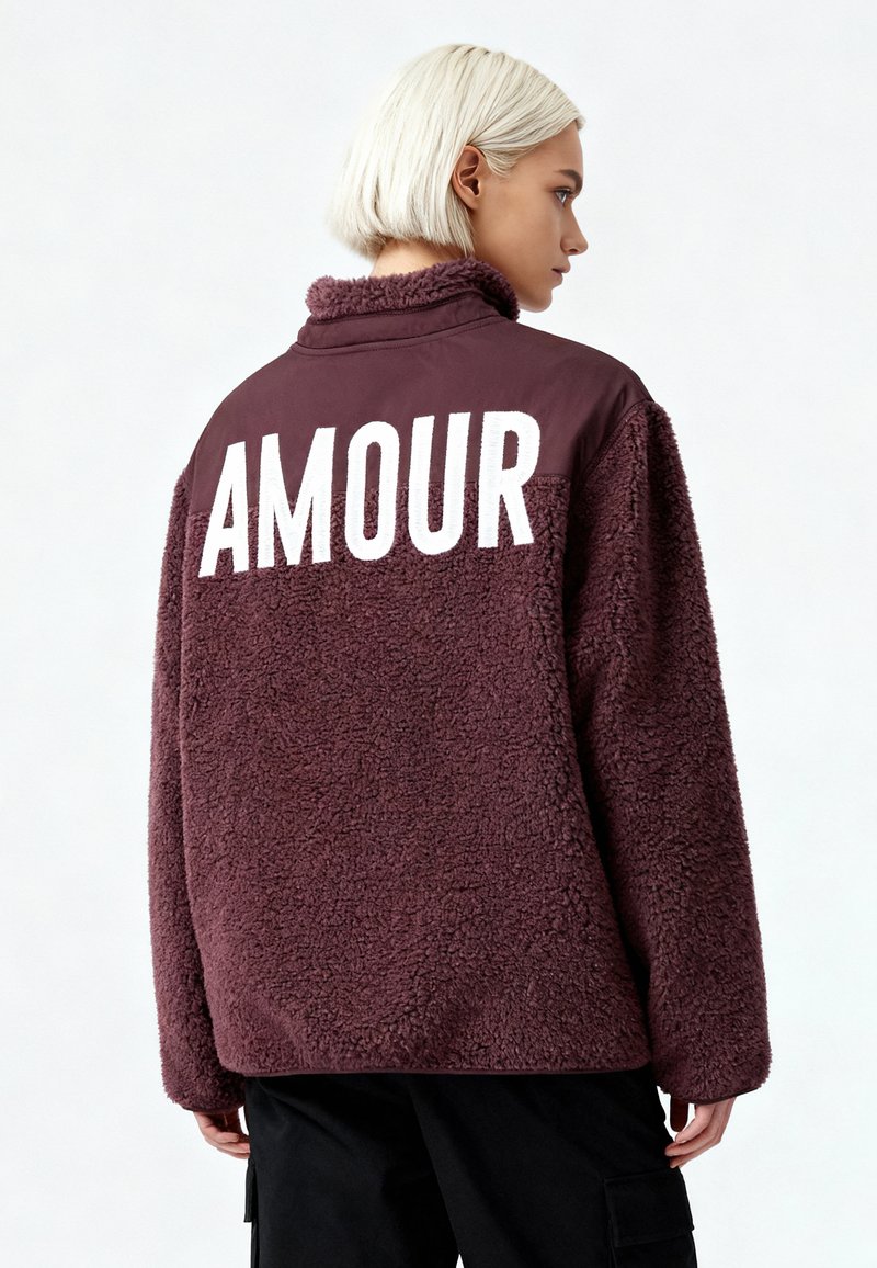 Veste en polaire bordeaux avec une finition texturée, dotée d'un col montant et de grandes lettres blanches "AMOUR" au dos.