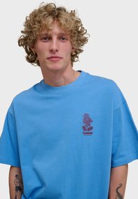 T-shirt en coton bleu clair avec un col rond et des manches courtes. Le devant affiche un logo graphique avec un personnage et le nom de la marque.
