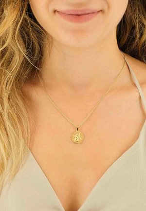 Collier pendentif en or avec un charme rond texturé présentant un design détaillé, suspendu à une fine chaîne en spirale. Porté sur un tissu clair.