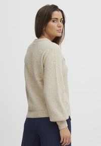 Beige neulepullover, jossa on tekstuurinen kangas, pallohihnat ja ribbaus hihansuissa ja helmassa. Pienen koristeelliset yksityiskohdat ovat näkyvissä edessä.