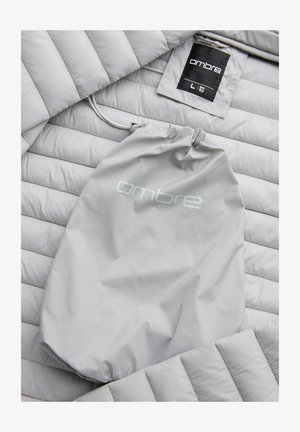 Giacca a vento grigia chiara con texture trapuntata e cappuccio regolabile. Accompagnata da una borsa di stoccaggio grigia abbinata con logo "ombre".