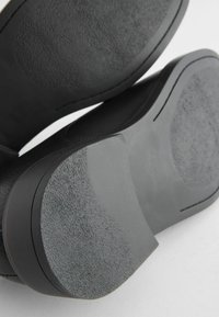 Zapatos negros con suelas de goma texturizadas, con una parte superior suave y bordes redondeados. La suela muestra texturas de superficie variadas.