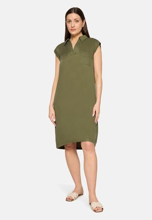 Femme aux longs cheveux foncés portant une robe chemise sans manches vert olive et des sandales plates beige, debout devant un fond uni.