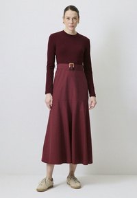 Lång burgundy klänning med åtsittande liv, långa ärmar och bälte i midjan. Utrustad med en utsvängd kjol i en slät textur; bärs med beige sneakers.
