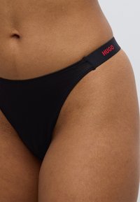 Μαύρο εσώρουχο στρινγκ με λεία υφή, διαθέτοντας ένα κόκκινο λογότυπο στη μέση. Απλός σχεδιασμός με ελάχιστη κάλυψη.