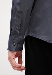 Σκούρο γκρι μακρυμάνικο πουκάμισο με κουμπωτές μανσέτες, με λεπτομέρειες σε αιχμηρές μανσέτες, λεία υφή υφάσματος και δύο κουμπι�ά σε κάθε μανσέτα.