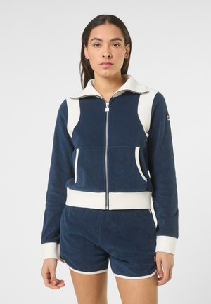 Donna con capelli lisci e scuri che indossa una giacca con zip blu navy e bianca e pantaloncini abbinati, su uno sfondo semplice.