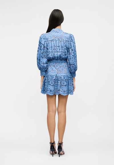 Woman with long dark hair wears a blue floral lace dress with puffed sleeves and black high-heeled sandals, standing facing away.