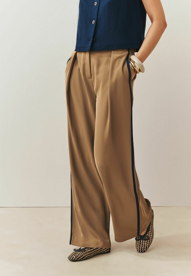 Personne portant un pantalon ample beige avec des bandes latérales noires, un haut sans manches bleu marine, des bracelets épais et des chaussures slip-on à motifs, debout les mains dans les poches.