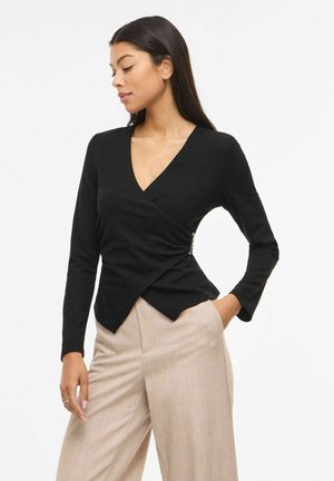 Žena v čiernom zavinovacom dlhom rukáve a béžových širokých nohaviciach pózuje s jednou rukou v vrecku na bielom pozadí.