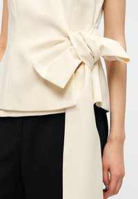 Κρεμ τοπ πεπλούμ από ελαφρύ ύφασμα, με μεγάλη φιόγκο στη μέση και ομαλή υφή. Συνδυάζεται με σκουρόχρωμο παντελόνι.