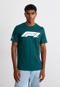 Grøn bomulds t-shirt med et stort hvidt Formel 1-logo på forsiden, korte ærmer og rund halsudskæring. Lyseblå jeans.
