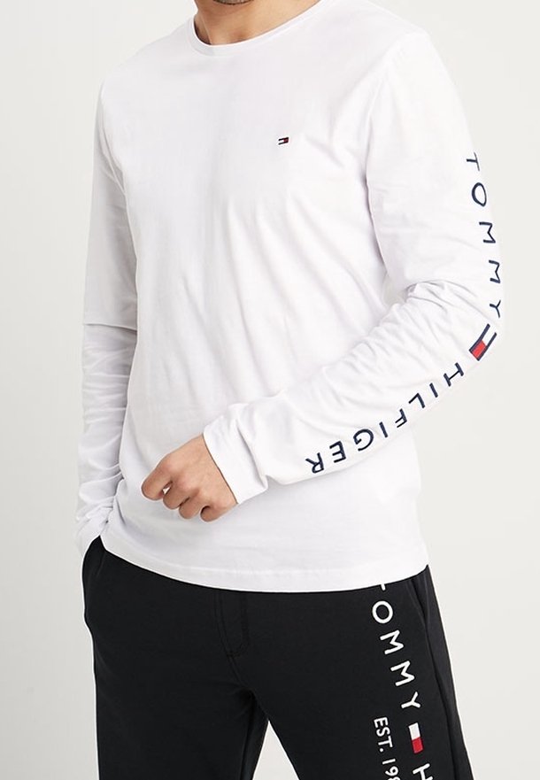 Man som bär en vit långärmad skjorta med "Tommy Hilfiger"-text och logotyp på ärmen, tillsammans med svarta byxor med matchande varumärke.