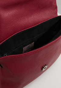 Červená texturovaná kožená taška s magnetickým zapínáním, vybavená černou látkovou vnitřní částí a vnitřní kapsou na zip. Uvnitř je viditelná etiketa.