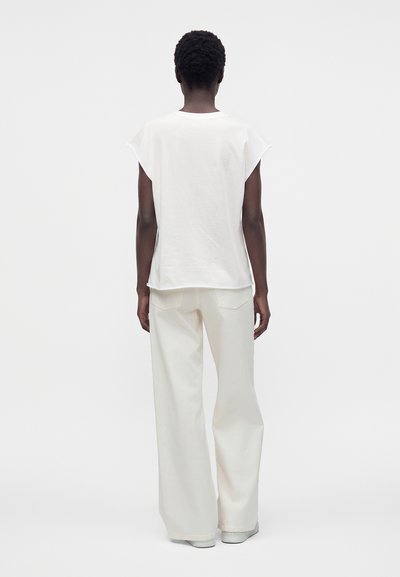 Personne aux cheveux courts et bouclés, vêtue d’un haut blanc ample sans manches et d’un pantalon blanc à jambes larges, debout de dos sur un fond uni.