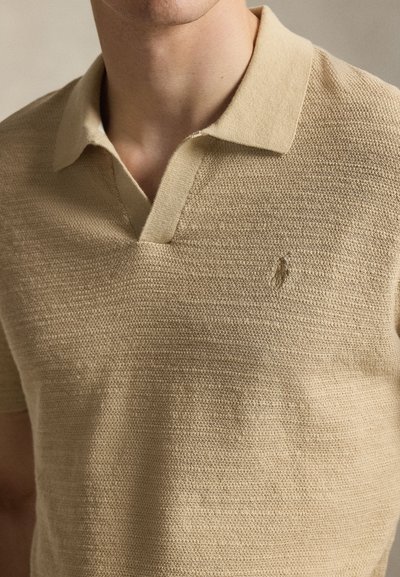 Μπεζ πλεκτό πόλο με V-λαιμόκοψη και κοντά μανίκια· διαθέτει διακριτικό υφασμάτινο σχέδιο και ένα μικρό κεντημένο λογότυπο στο στήθος.