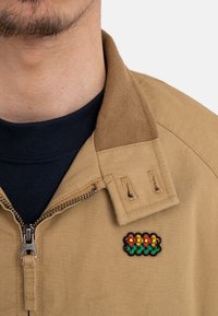 Tan zipped jacket with a textured collar, featuring a small embroidered floral patch in red, yellow, and green on the left side.