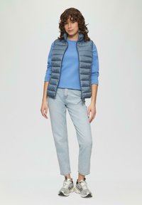 Blaues Steppweste über einem hellblauen Langarmshirt, kombiniert mit hellblauen Jeans und silbernen Sneakers. Weicher Stoff und lässiges Design.