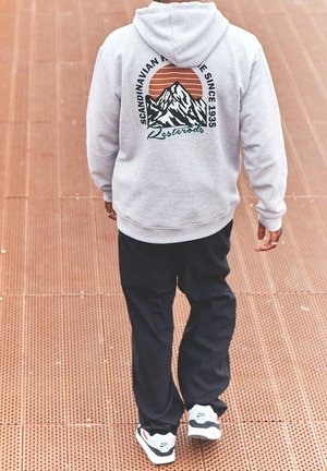 Grijze hoodie met bergafbeelding, oranje zon en tekst. Gecombineerd met zwarte broek en witte sneakers met rode en groene accenten.