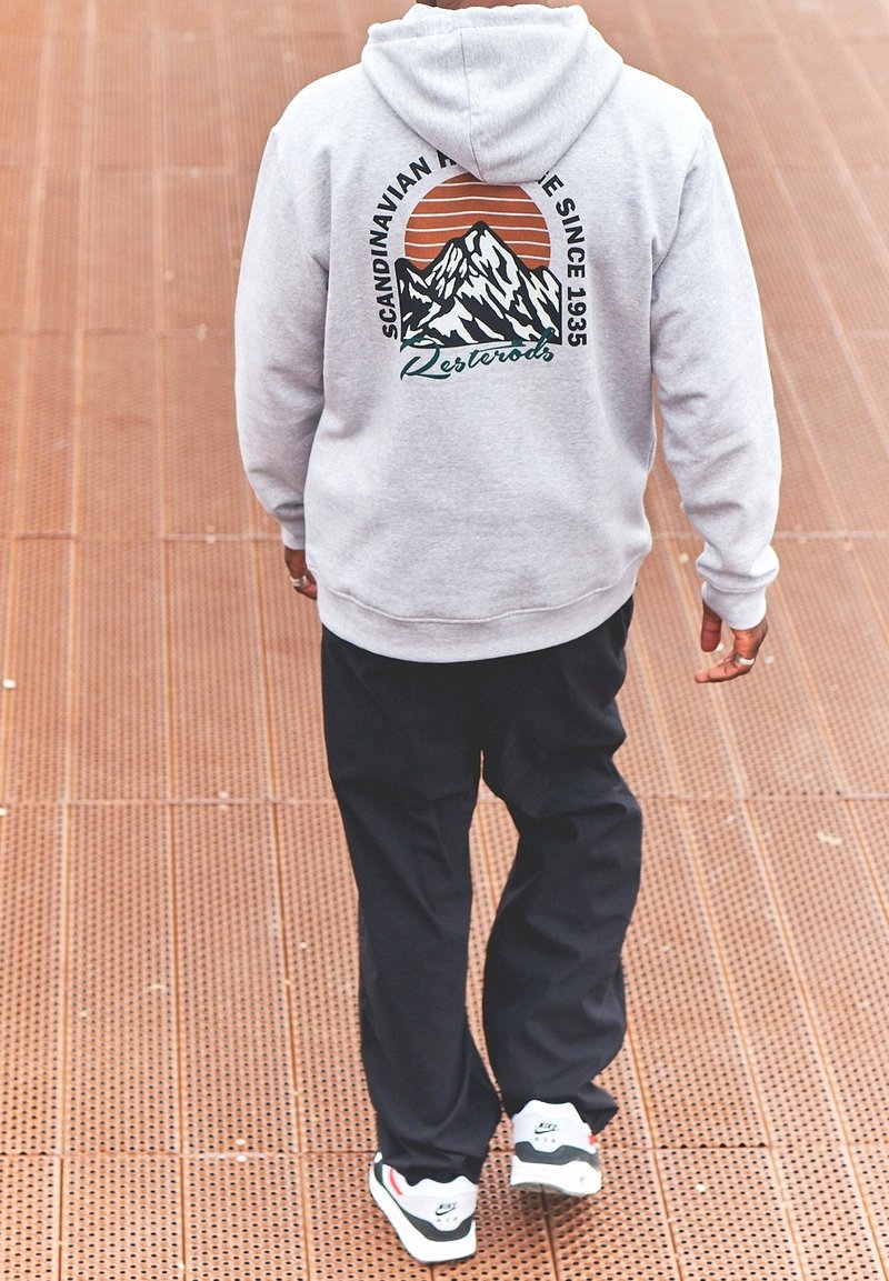 Harmaa huppari, jossa on vuori graafinen kuvasto, oranssi aurinko ja teksti. Yhdistetty mustiin housuihin ja valkoisiin tennareihin, joissa on punaisia ja vihreitä yksityiskohtia.