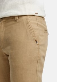 Pantalones de algodón beige con bolsillos laterales, corte ajustado y una textura suave, adornados con un pequeño detalle de costura multicolor.