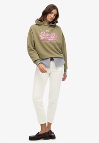 Felpa khaki con testo grafico rosa, indossata sopra una camicia a righe blu. Abbinata a jeans bianchi a gamba dritta e mocassini scuri.