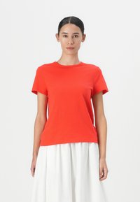 Tricou roșu din bumbac cu mâneci scurte, decolteu rotund și o croială lejeră. Purtați cu o fustă plisată albă, pe un fundal simplu.