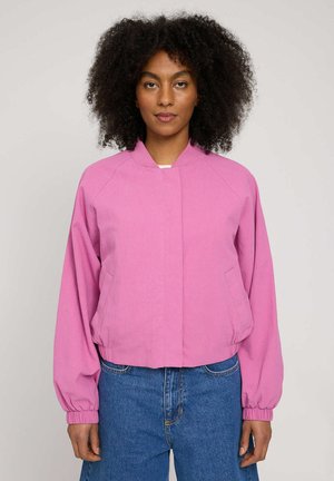 Woman with curly hair wearing a pink bomber jacket and blue jeans, standing facing forward against a neutral background.