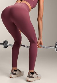 Persona che indossa leggings da allenamento viola e scarpe da ginnastica solleva un bilanciere con pesi in un ambiente interno neutro.