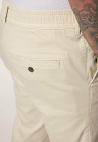 Beige broek met een elastische tailleband, met een achterzak met een bruine knop en subtiele stiksels. Soepele textuur.