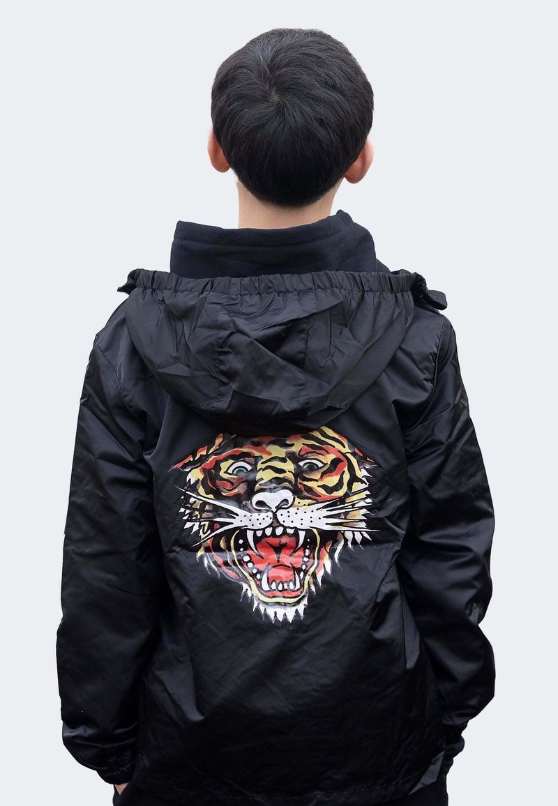 Personne aux cheveux courts et foncés portant une veste noire avec un grand dessin de visage de tigre rugissant dans le dos, debout de dos.