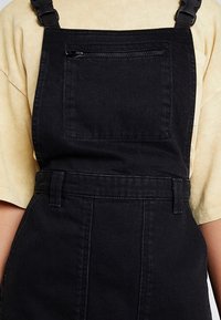 Svarta denimoverall med framficka med dragkedja, bärs över en beige kortärmad skjorta, visar torso och övre lår.