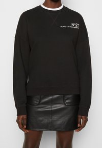 Sudadera de felpa negra con puños acanalados, que presenta el texto blanco "N°21 MILANO - VIA SANTO SPIRITO" en el pecho. Combinada con una falda corta de cuero negra.