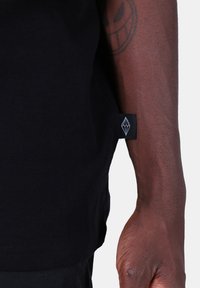 Černé bavlněné tričko s božní etiketou a bílým geometrickým logem. Tkanina vypadá hladce a má běžný střih.