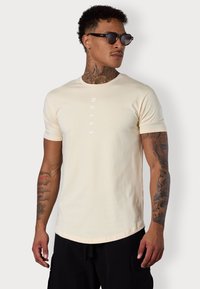 Beige T-Shirt mit kurzen Ärmeln, Rundhalsausschnitt und weißem, vertikalem Textdesign. Der Stoff wirkt glatt und hat eine taillierte Silhouette.