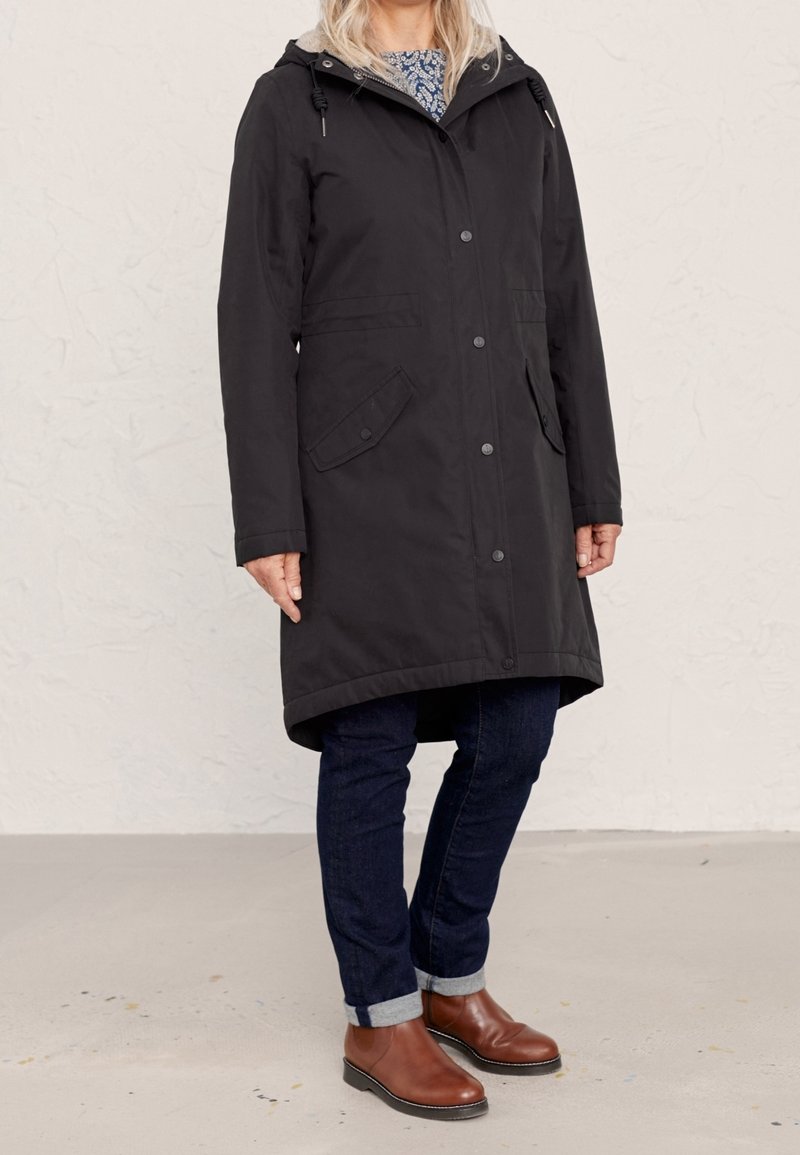Czarna wodoodporna parka z kapturem, zapięciem na napy z przodu, kieszeniami bocznymi i dłuższym tyłem. Nosi pod spodem ciemne dżinsy i brązowe botki za kostkę.