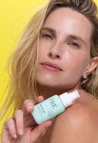 Jasno miętowa butelka do dozowania z etykietą "ODŻYW MNIE", z białą zakrętką. Modelka z mokrymi, falującymi blond włosami, na tle jasnego żółtego koloru.