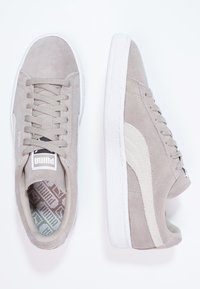 Helle graue Wildleder-Sneaker mit weißen Gummisohlen, Schnürverschluss und einem beigen Streifen an der Seite. Mit Logo-Patch auf der Zunge.