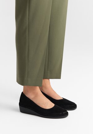 Black suede wedge shoes with a rounded toe and low heel, worn with olive green tailored pants that have a straight cut and side slits.