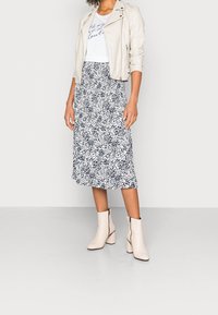 Chaqueta de motorista de cuero blanco sobre una camiseta gráfica gris claro, combinada con una falda larga con manchas en blanco y negro y botas de tobillo crema.