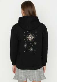 Sudadera con capucha negra que presenta un diseño decorativo en la parte posterior con patrones de estallido de estrellas y adornos. La tela parece suave y texturizada.