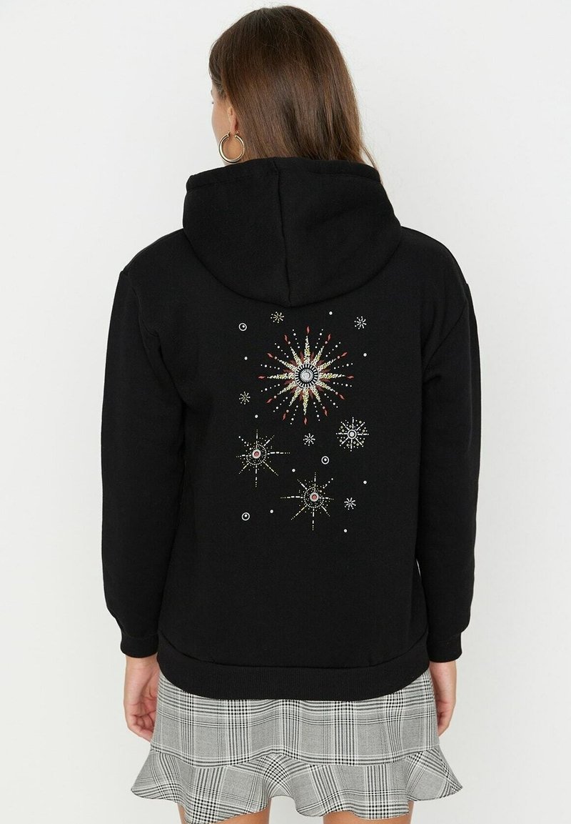Sudadera con capucha negra que presenta un diseño decorativo en la parte posterior con patrones de estallido de estrellas y adornos. La tela parece suave y texturizada.