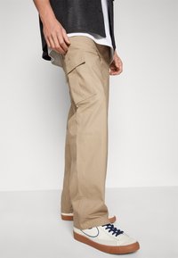 Beige cargo bukser med sidelommer, en løs pasform og subtil tekstur; parret med hvide sneakers med marineblå snørebånd og gummisåler.