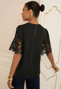 Blusa nera con maniche corte in pizzo, caratterizzata da un collo rotondo e una scollatura a chiave sul retro. Tessuto liscio con dettagli in pizzo intricato sulle maniche.
