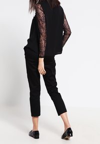 Outfit nero composto da un top in pizzo trasparente con maniche lunghe e pantaloni neri su misura. Il top ha una vestibilità comoda, con retro fluido e maniche aderenti.