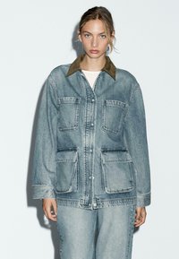 Denimjack met een bruine kraag, twee grote voorzakken en een ontspannen pasvorm. Lichtblauwe wassing met zichtbare stikseldetails.