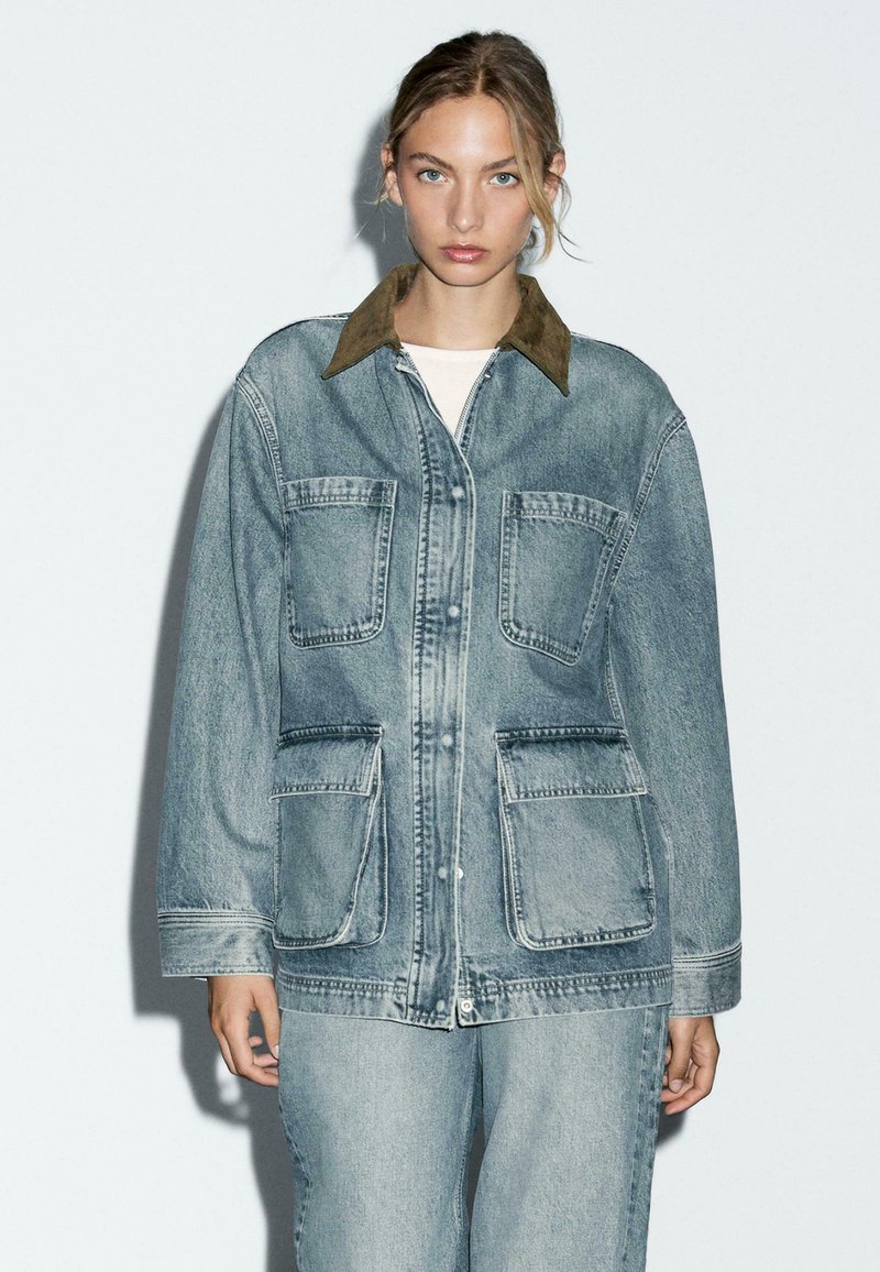 Denimjack met een bruine kraag, twee grote voorzakken en een ontspannen pasvorm. Lichtblauwe wassing met zichtbare stikseldetails.