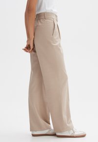 Beige wijde broek met geplooide voorkant, elastische tailleband en achterzakken, gecombineerd met witte sneakers en een eenvoudig t-shirt.